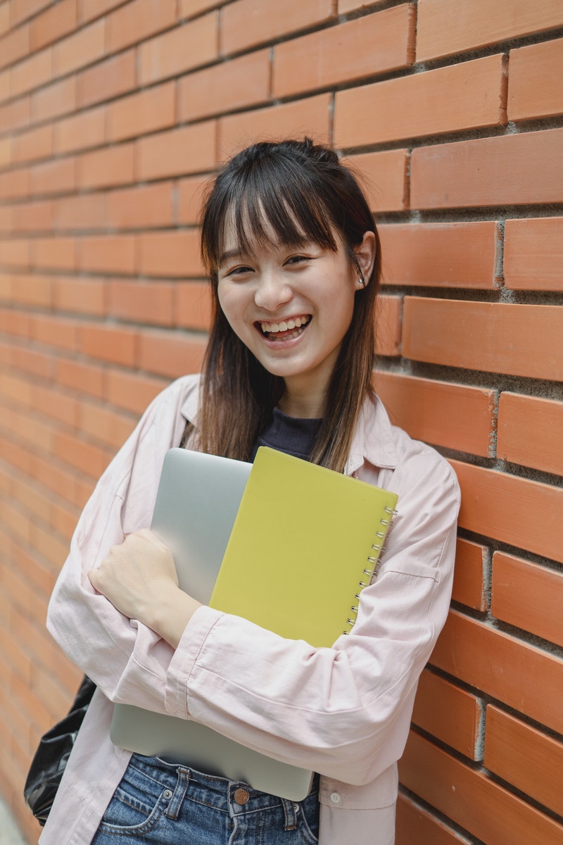 Joyful Asian female student leaning on university brick wall